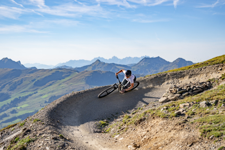 Bike fährt in einer Kurve den Bike-Trail hinunter, im Hintergrund sieht man die Berglandschaft
