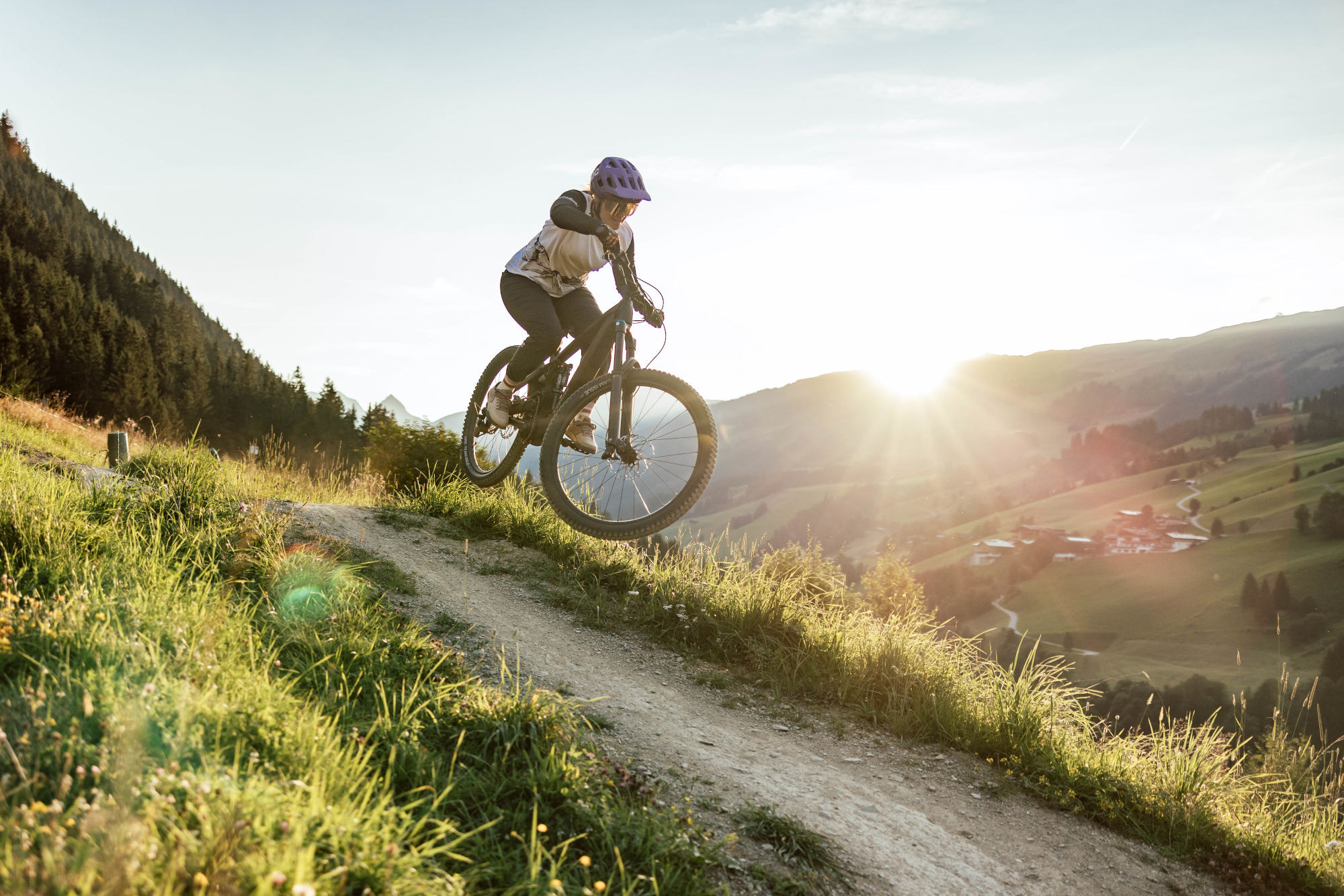 Cyclist wearing a helmet rides a mountain bike on a grassy trail, catching air against a backdrop of rolling hills and a setting sun. Adventure and freedom.
