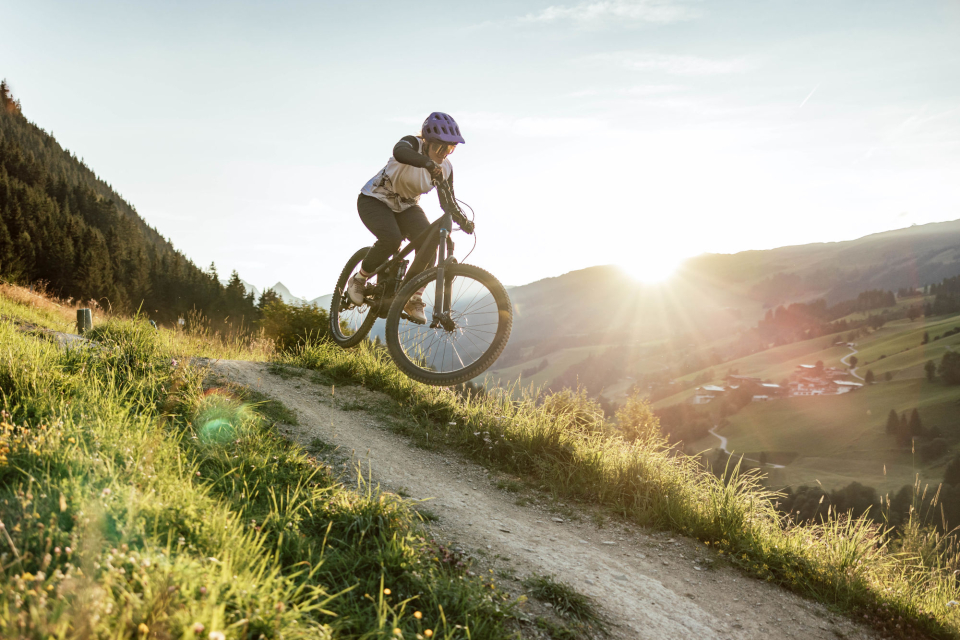 Ein Radfahrer mit Helm fährt mit seinem Mountainbike auf einem grasbewachsenen Weg und springt vor dem Hintergrund sanfter Hügel und der untergehenden Sonne in die Luft. Abenteuer und Freiheit.