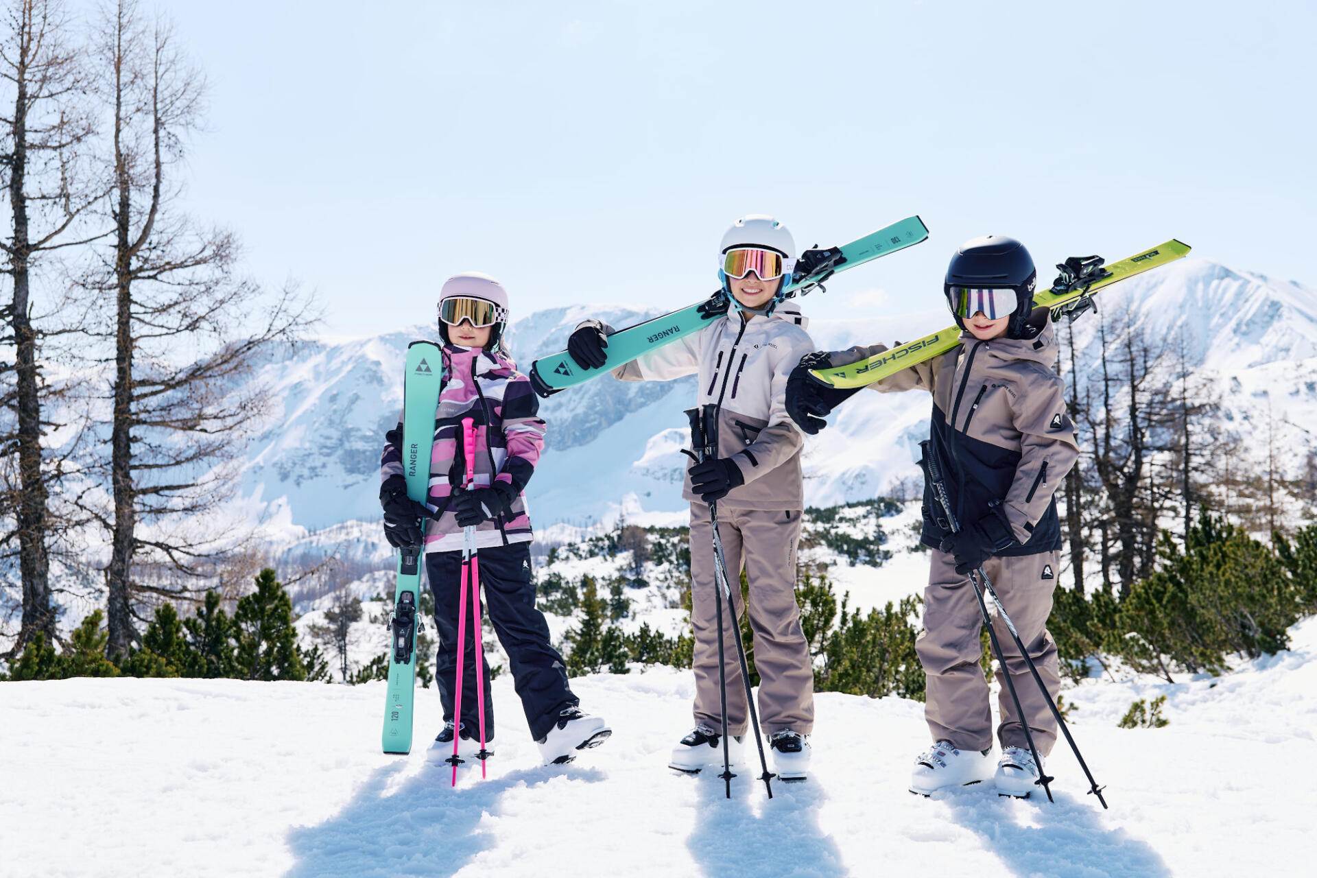 Three children standing in the snow holding their colourful skis in their hands or carrying them on their shoulders