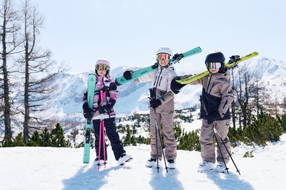3 Kinder stehen im Schnee und tragen halten ihre bunten Skier in der Hand bzw. tragen sie auf der Schulter