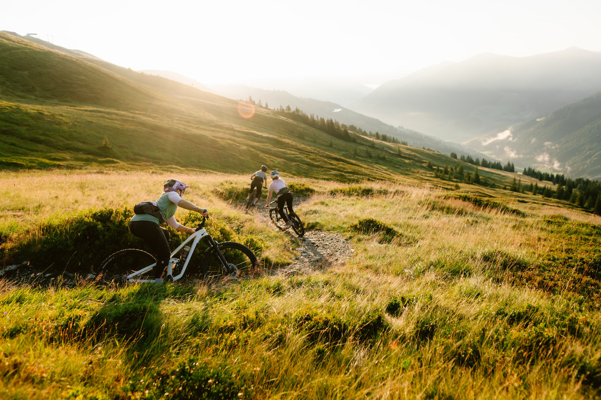 3 Biker fahren einen Bike Trail hinunter. Es ist Sonnenuntergang, dadurch erscheint die Landschaft in einem hellen und warmen Licht