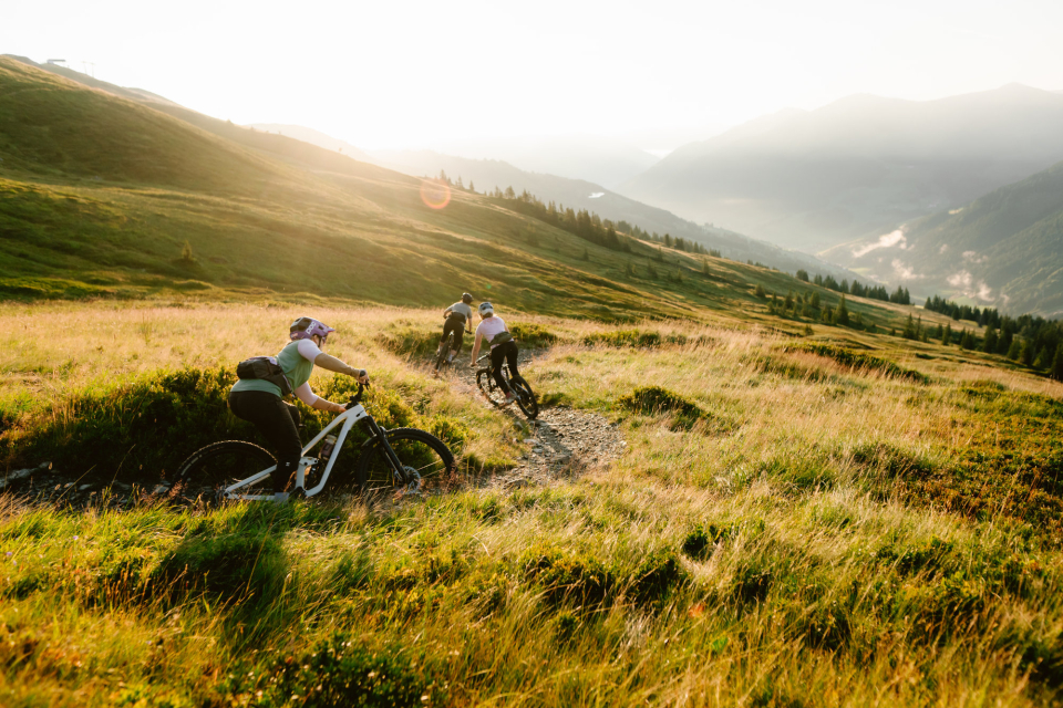 3 Biker fahren einen Bike Trail hinunter. Es ist Sonnenuntergang, dadurch erscheint die Landschaft in einem hellen und warmen Licht