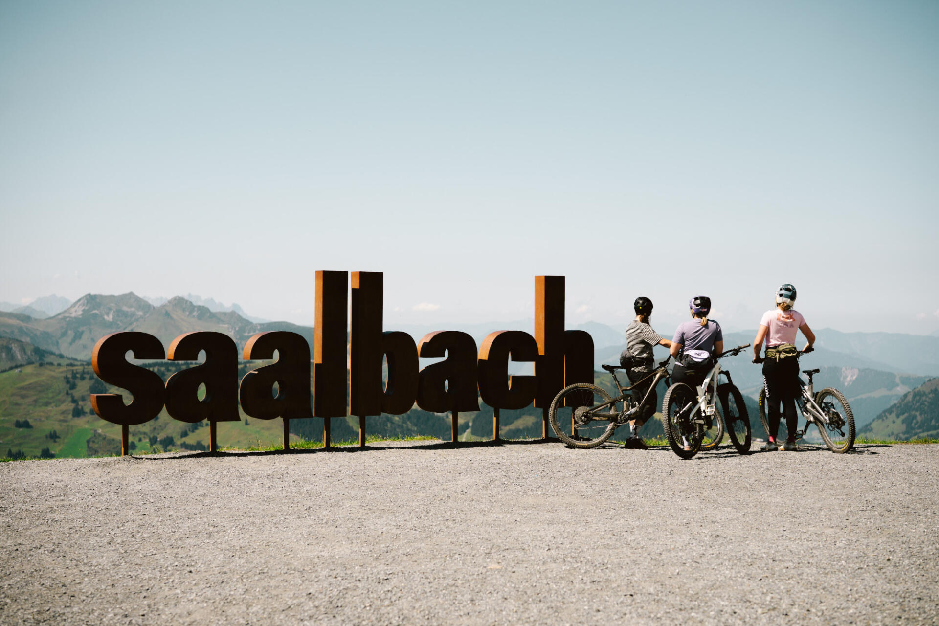 Three bikers stand next to a sculpture consisting of the words "Saalbach" and look out over the landscape.