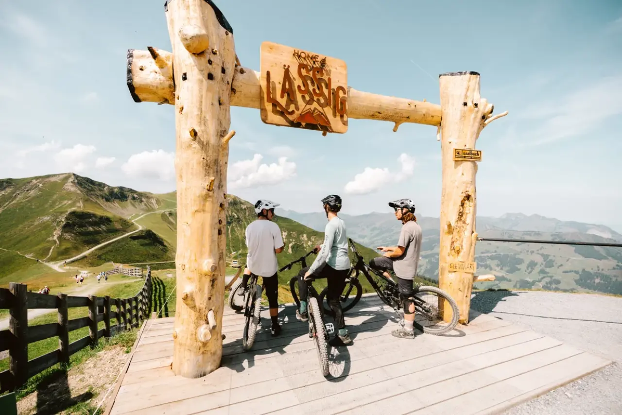Three cyclists stand under a Home Of Lässig gate and look out at the mountain landscape.