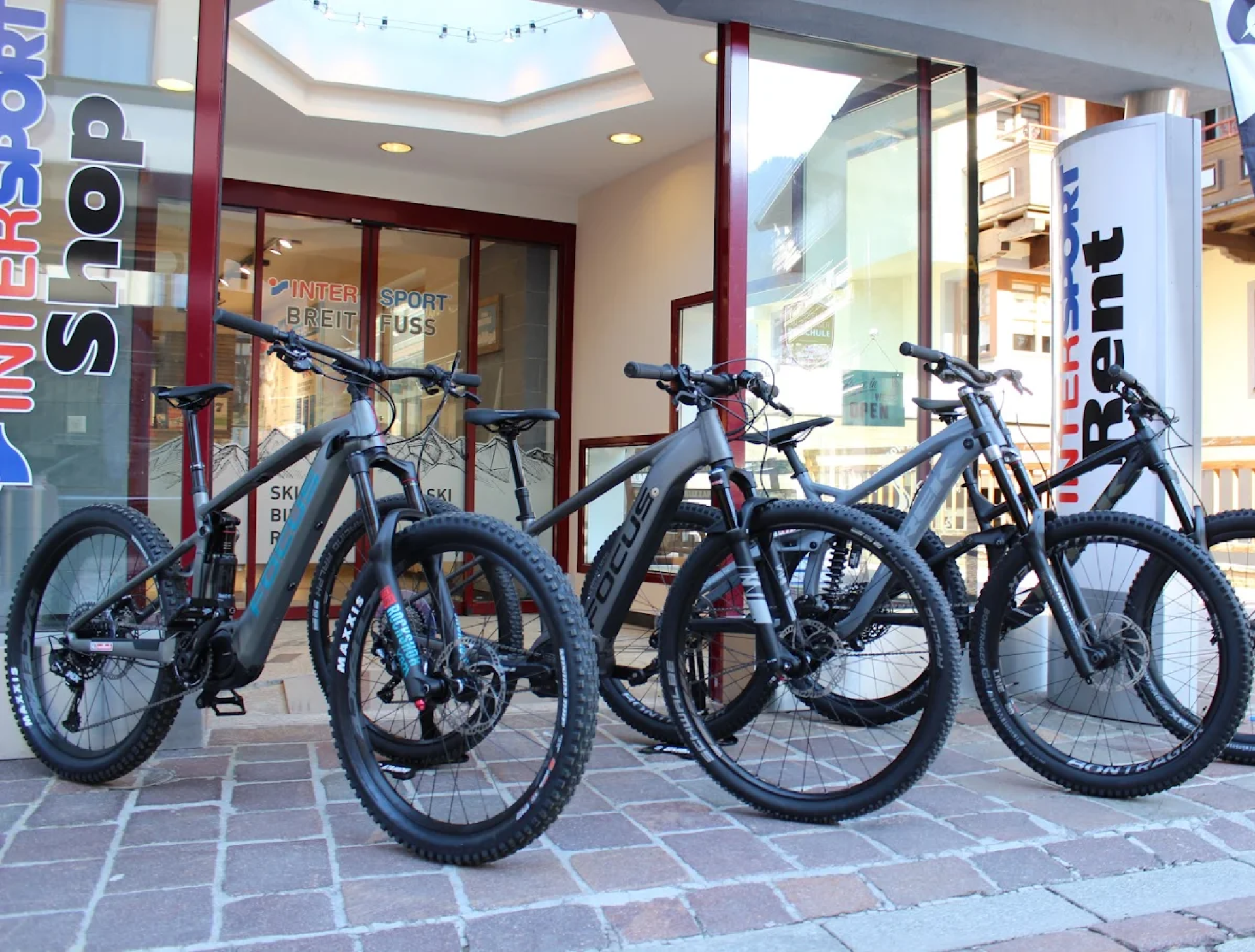 Vier Mountainbikes mit dicken Reifen stehen vor einem Intersport-Verleihgeschäft auf einem Kopfsteinpflasterweg in einer Reihe. Das Geschäft hat Glasfenster und eine Tür mit rotem Rahmen.