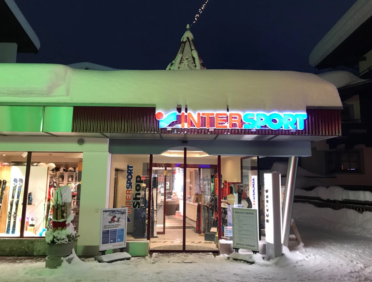 Das Bild zeigt einen gut beleuchteten Intersport-Laden bei Nacht, der mit Schnee bedeckt ist. Die hellen Leuchtreklamen verleihen der winterlichen Kulisse Wärme und schaffen eine einladende Atmosphäre.