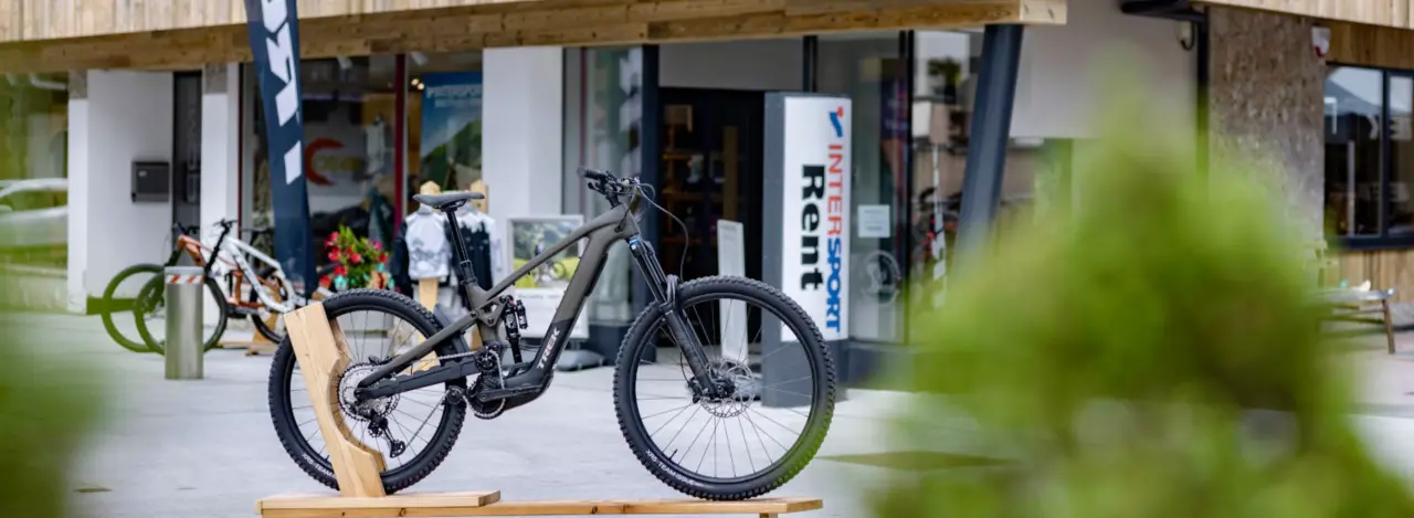 A sleek, black mountain bike displayed outdoors on a wooden stand. Behind it is a store with a glass front and a sign reading "Intersport Rent."
