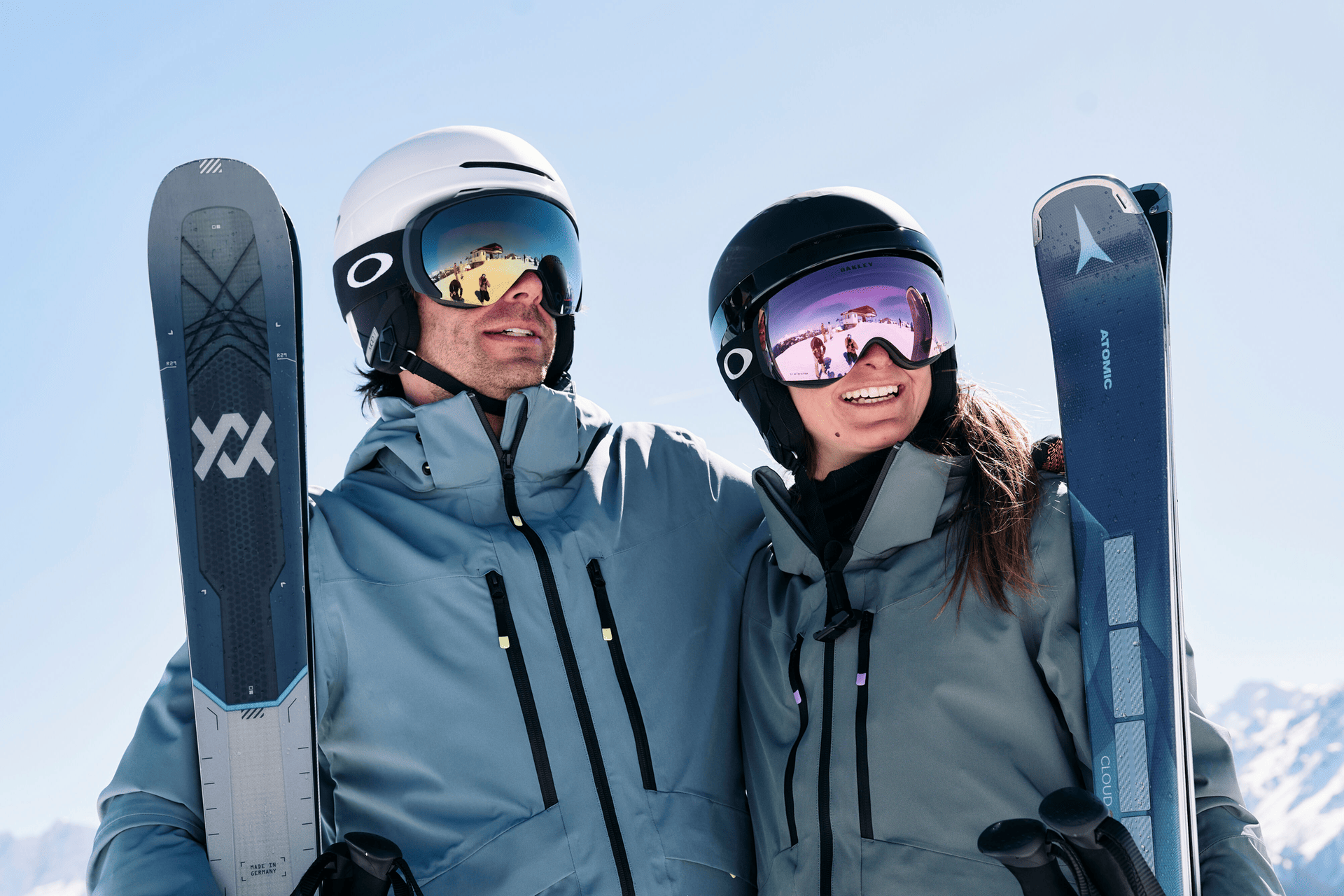 Three skiers in colorful gear joyfully raise their poles on a snowy mountain with a backdrop of majestic, sunlit peaks and a clear blue sky.
