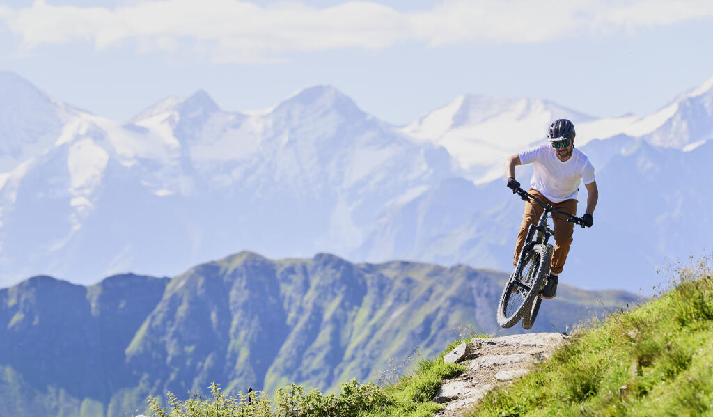Ein Radfahrer mit Helm und Schutzkleidung steht mit seinem Mountainbike auf felsigem Gelände und blickt unter einem klaren Himmel auf eine ruhige Berglandschaft.