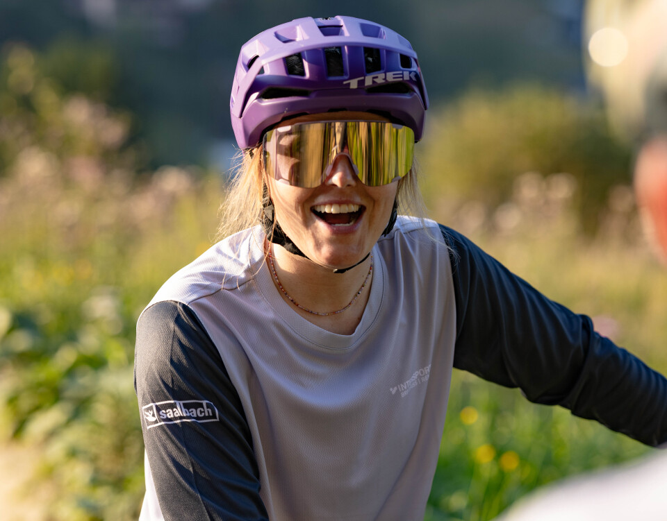 Eine Bikerin lächelt in die Kamera, hinter ihr ist eine Wiesen-Landschaft zu sehen.