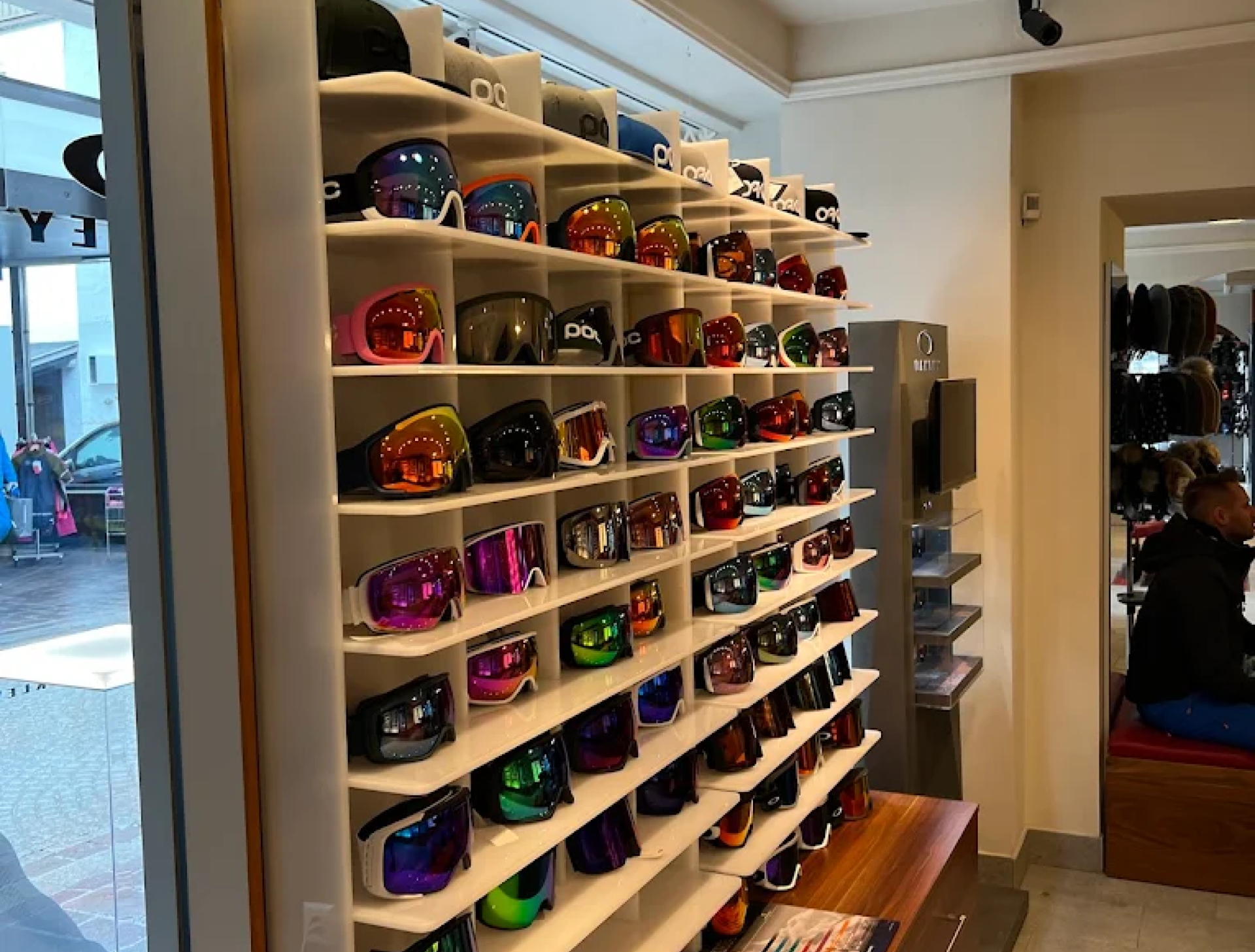 An einer Wand hängen bunte Ski- und Snowboardbrillen in Regalen in einem gut beleuchteten Laden. Eine Person sitzt auf einer Bank in der Nähe und sorgt für eine lebe