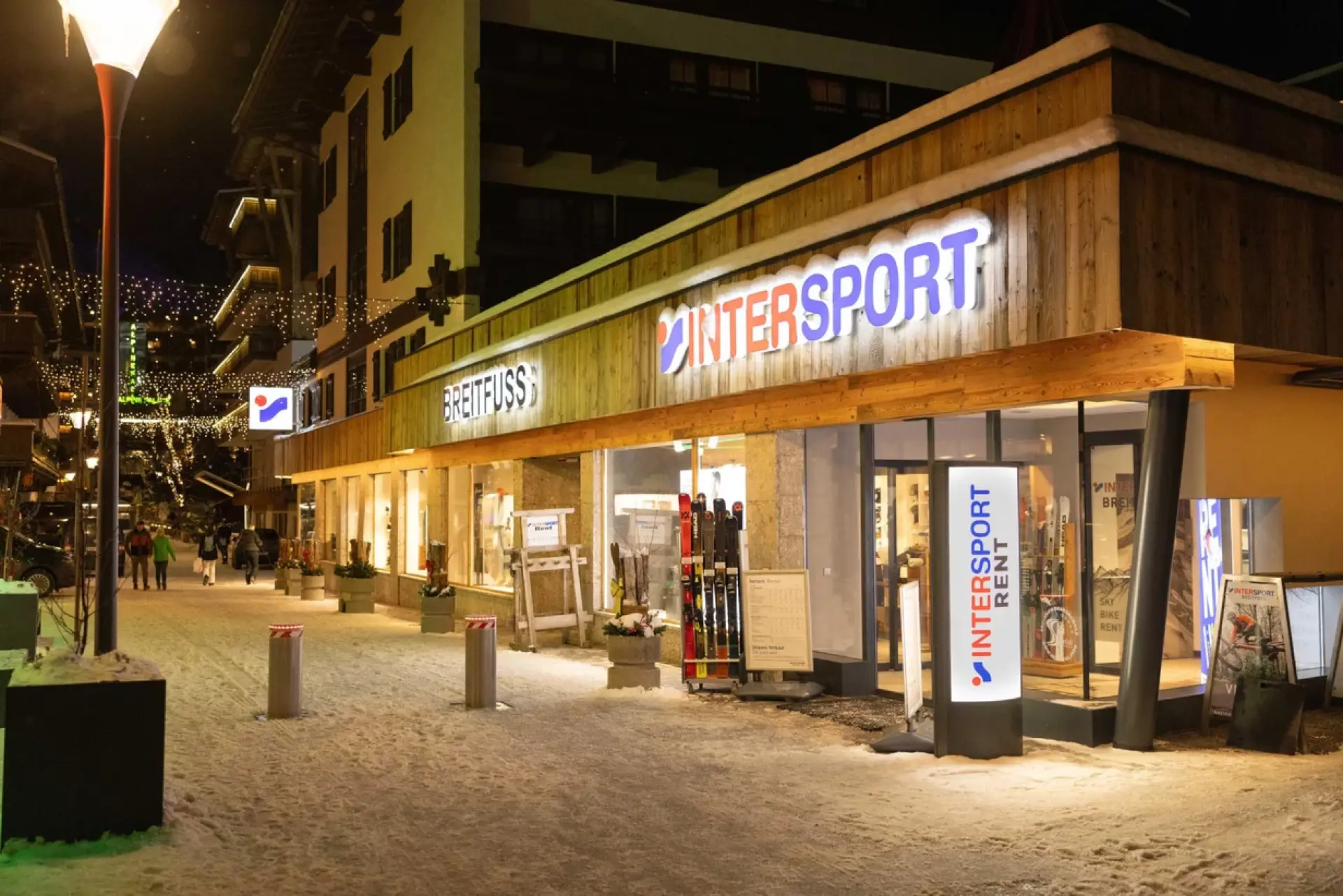 Nighttime view of a well-lit sports store, Intersport, on a snowy street. The warm lights create a welcoming ambiance with a few people walking by.