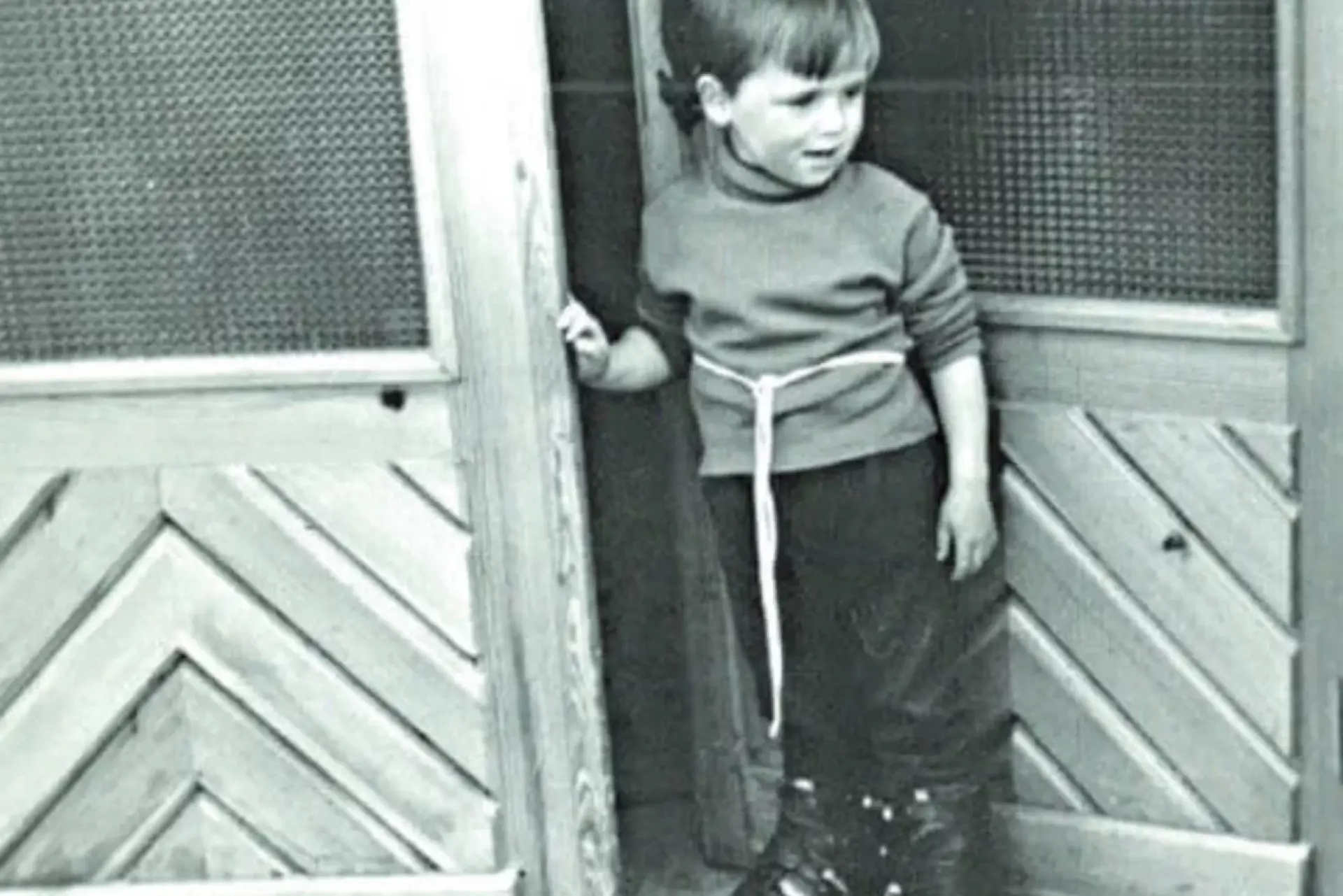 A young child in a sweater and pants stands in front of a wooden door. The child is looking to the side with a curious expression. The setting is retro.