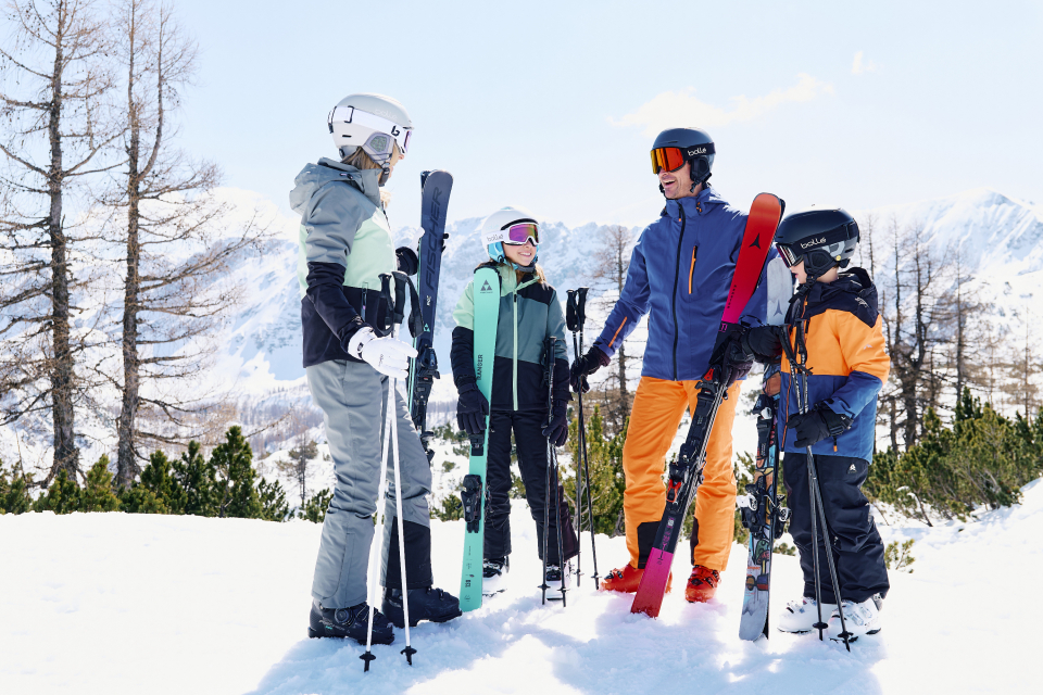 Eine vierköpfige Familie steht auf einem schneebedeckten Berggipfel, bekleidet mit Skiausrüstung und Helmen. Im Hintergrund sind schneebedeckte Berge und ein klarer blauer Himmel zu sehen.