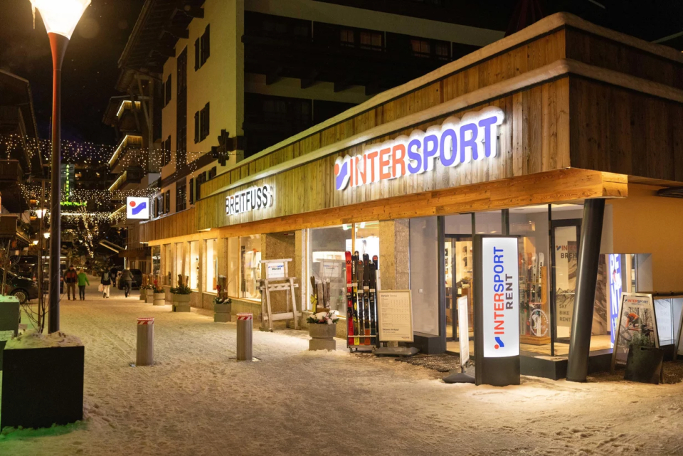 Nachtansicht eines hell erleuchteten Sportgeschäfts, Intersport, in einer verschneiten Straße. Die warmen Lichter schaffen eine einladende Atmosphäre, während einige wenige Passanten vorbeigehen.