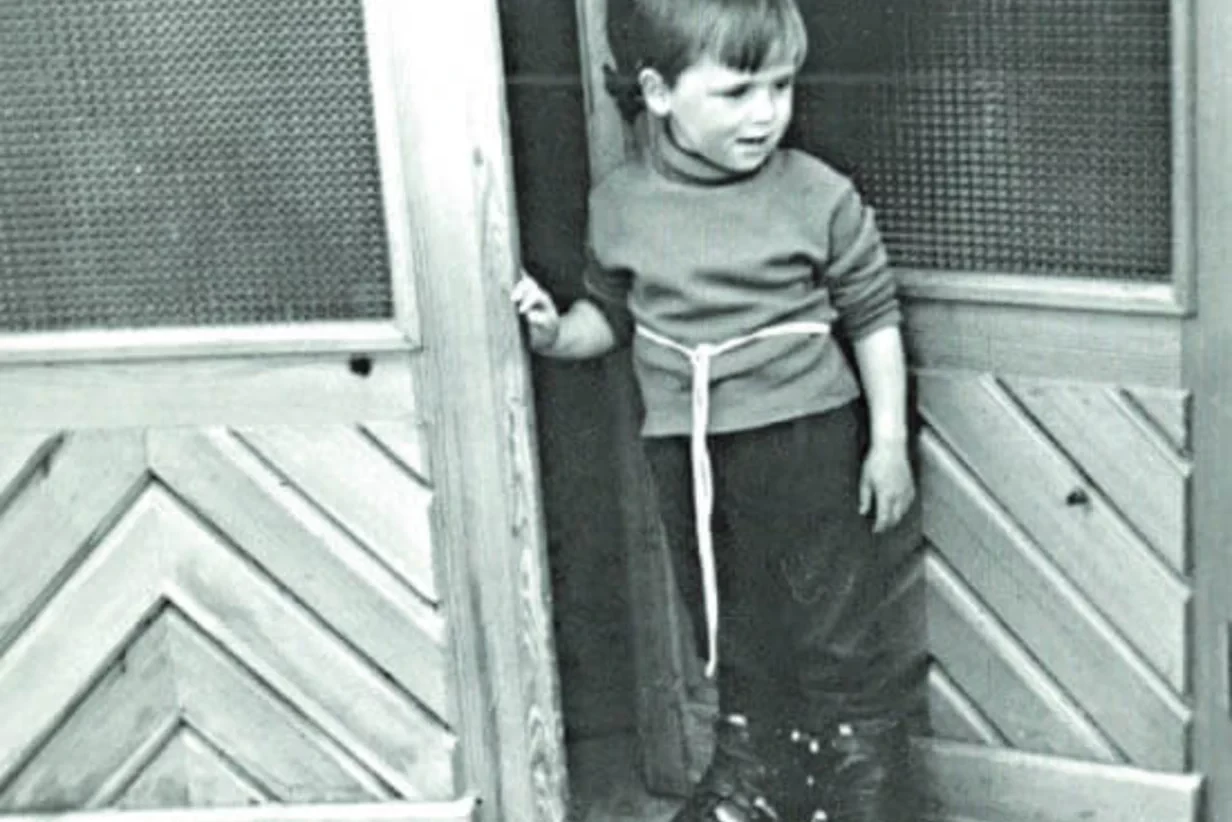 Ein kleines Kind in Pullover und Hose steht vor einer Holztür. Das Kind schaut mit neugierigem Blick zur Seite. Die Kulisse ist im Retro-Stil gehalten.
