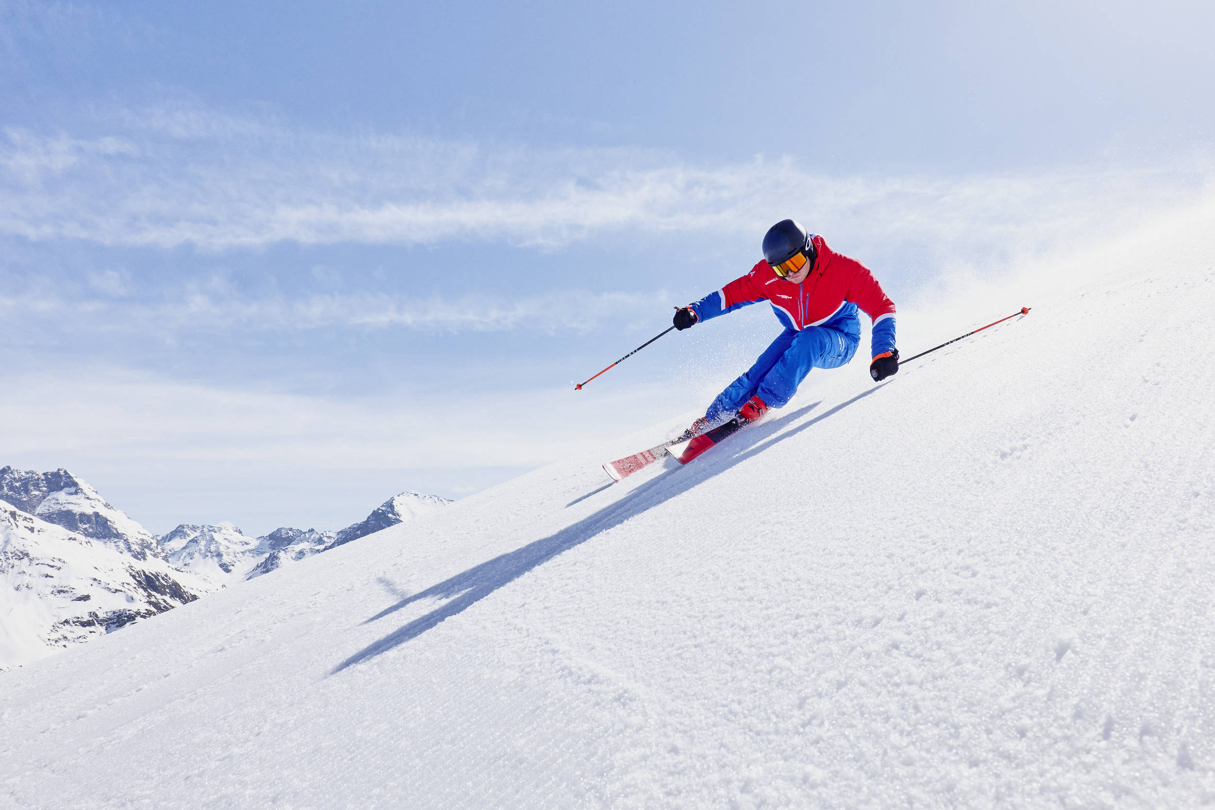 A skier in a red and blue outfit swiftly descends a snowy slope, wearing a helmet and goggles. Snowy mountains and a clear sky are in the background.