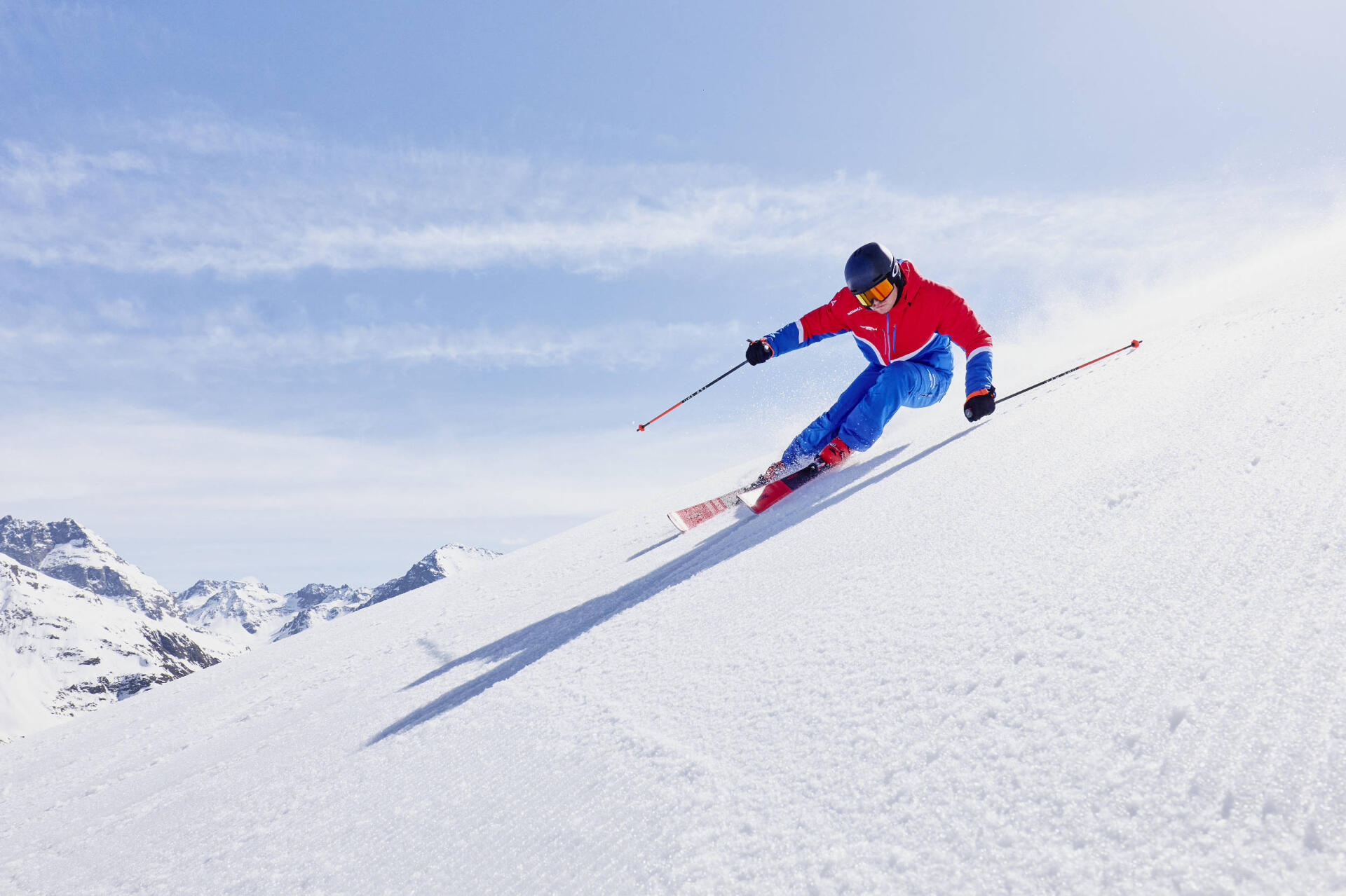 A skier in a red and blue outfit swiftly descends a snowy slope, wearing a helmet and goggles. Snowy mountains and a clear sky are in the background.
