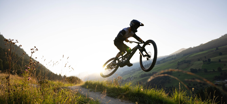 Mountainbiker springt bei Sonnenuntergang über einen Trail in den Bergen und fährt mit hoher Geschwindigkeit durch eine alpine Landschaft